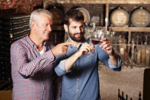 Portrait young sommelier standing at family wine cellar with senior winemaker and tasting a glass of red wine. Small business.
